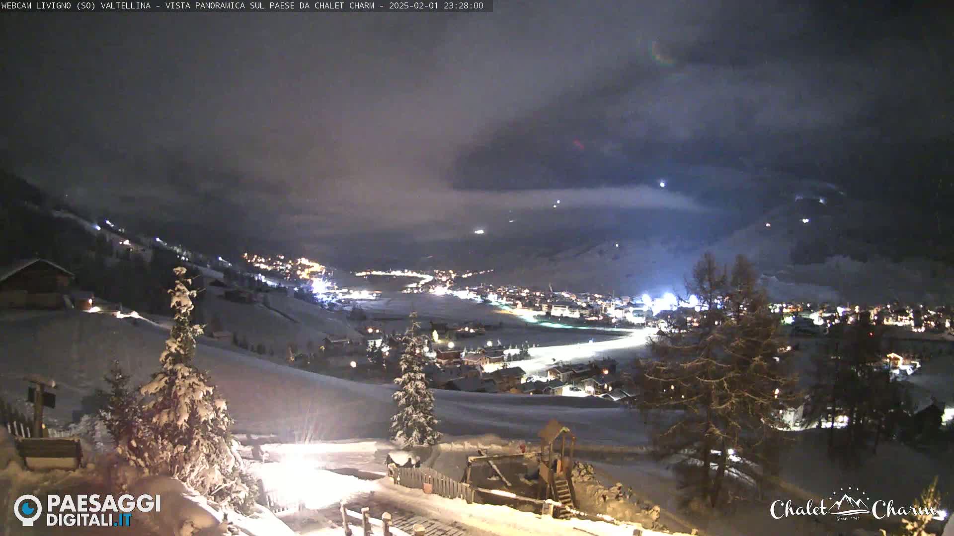 A snow-covered, nighttime view of a village nestled in a valley, under a cloudy sky.
