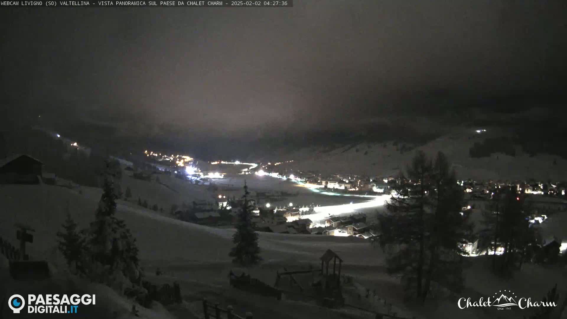 A snow-covered valley town is illuminated at night under a dark, cloudy sky.