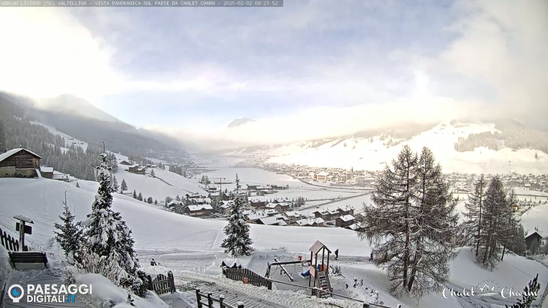 A snow-covered village nestled in a valley, with mountains in the background under a partly cloudy sky.
