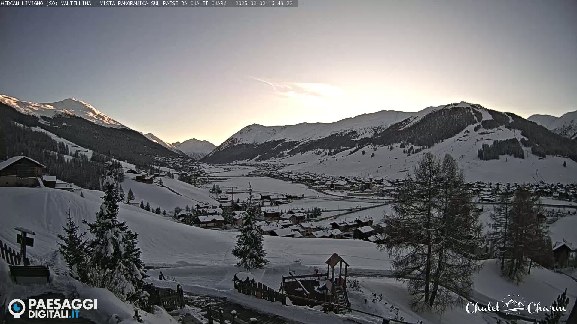 A snow-covered mountain village is nestled in a valley at sunset, under a clear, pale sky.