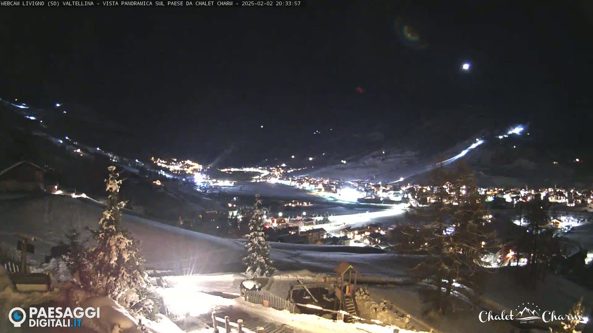 A nighttime, snow-covered mountain village is illuminated, with a clear sky showing a few stars and the moon.