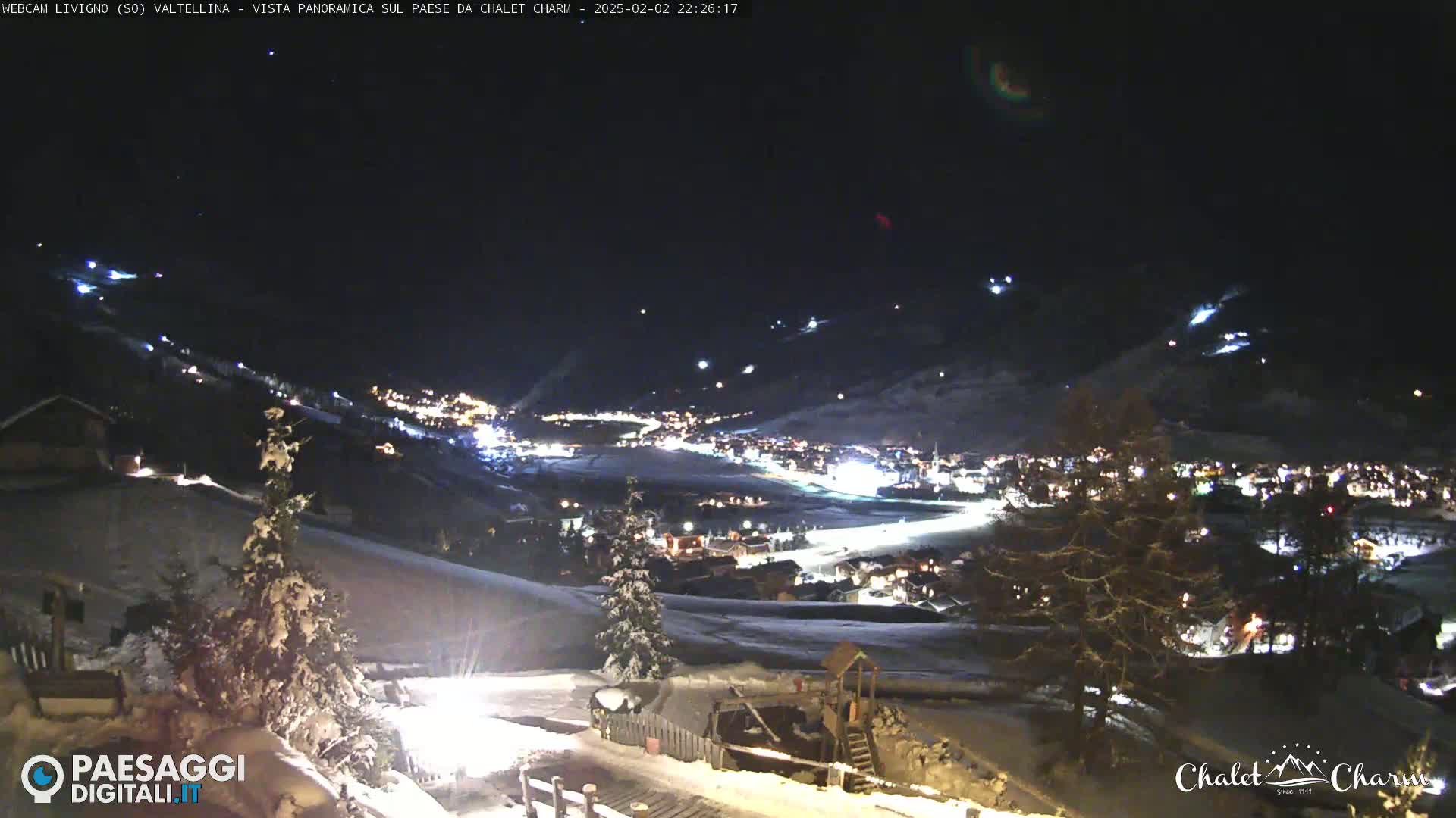 A snow-covered hillside at night overlooks a village illuminated by lights, under a dark sky.