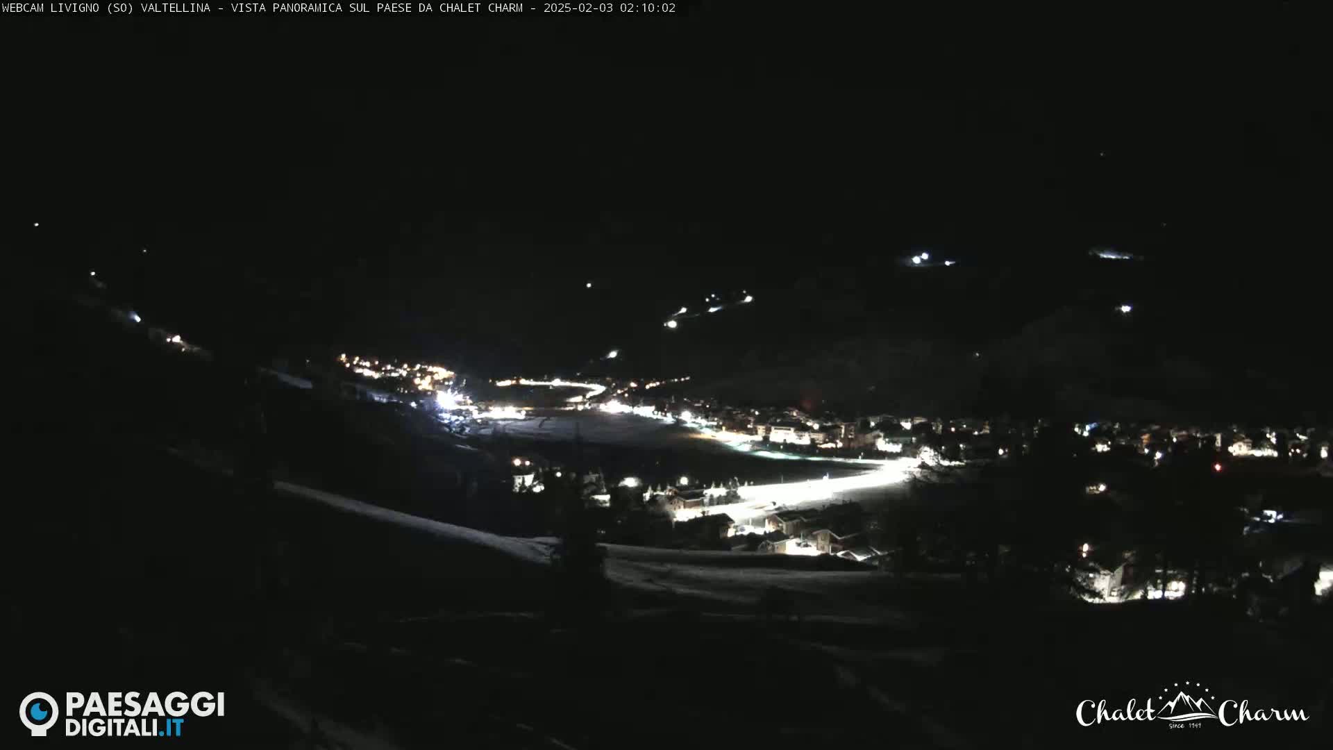 A nighttime panoramic view of a snow-covered town illuminated by lights, seen from a higher vantage point.