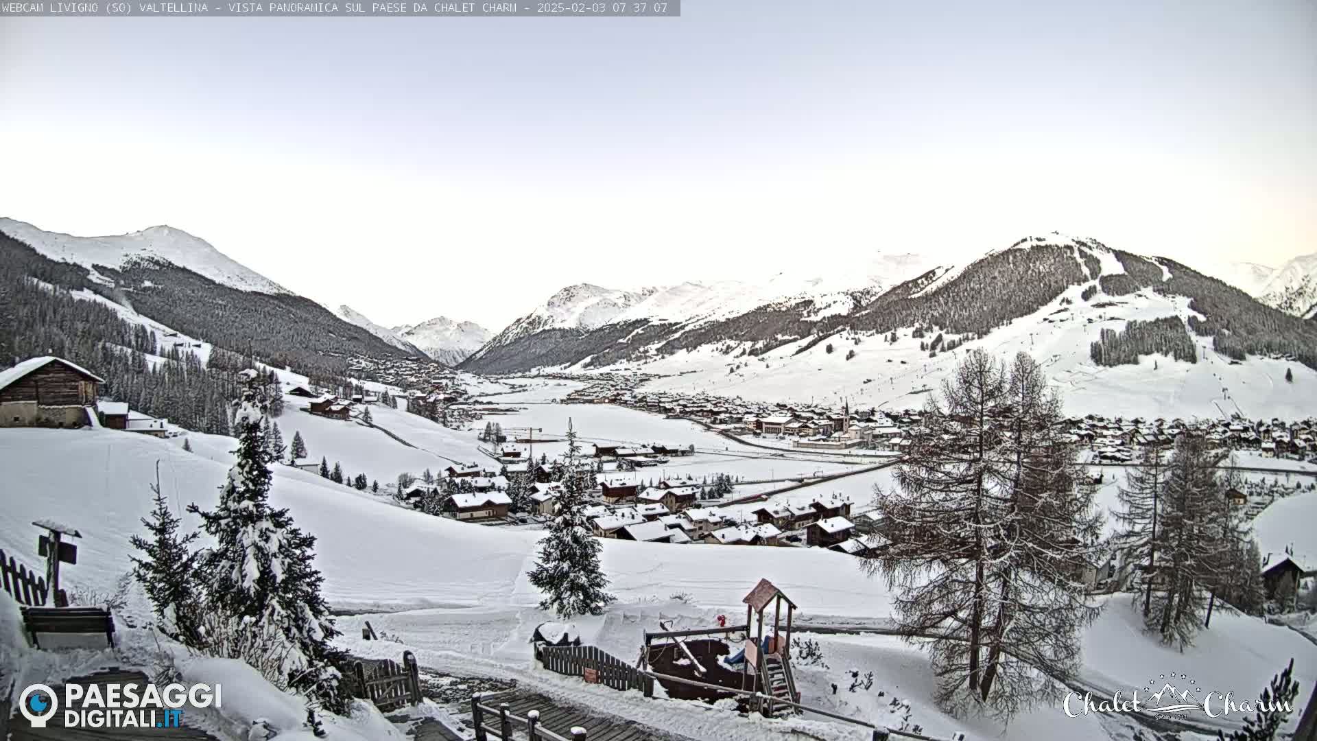 A snow-covered village nestled in a valley between snow-capped mountains under a clear, light sky.