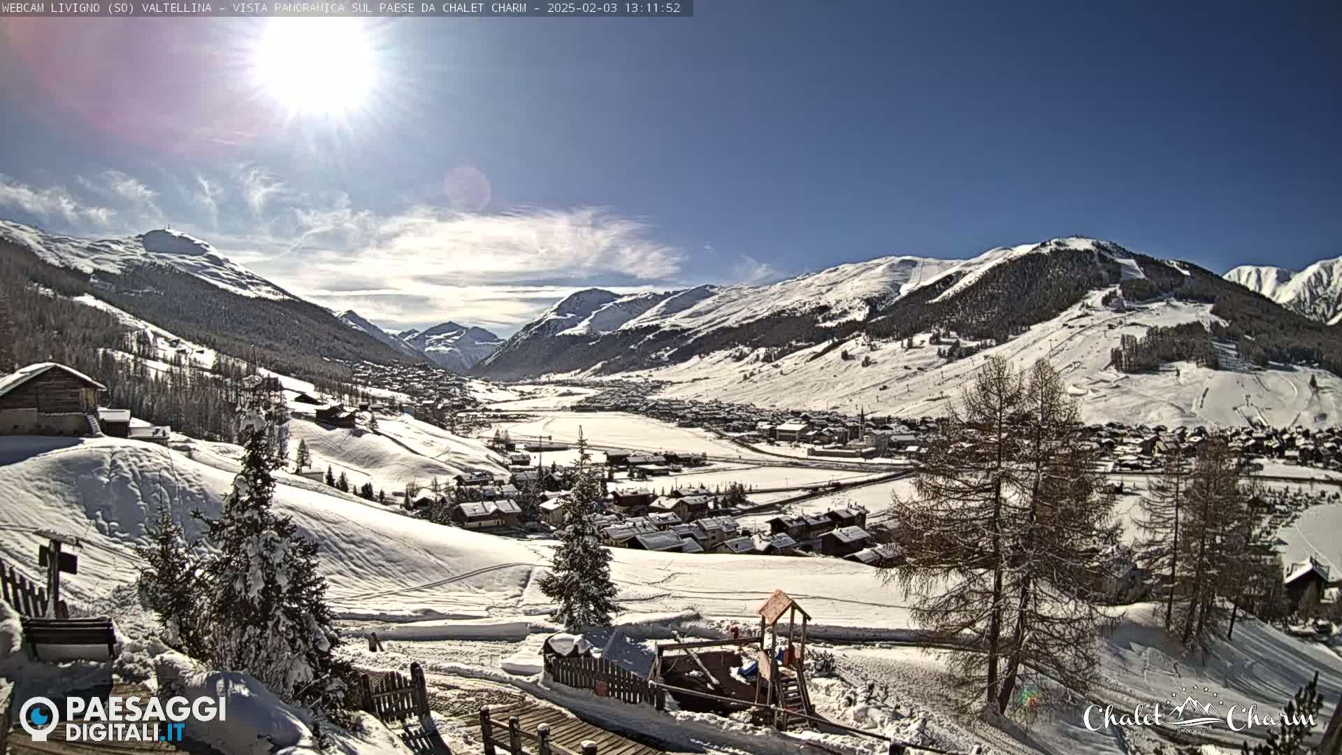 A sunny, snow-covered mountain valley village is seen from a hillside, with a playground visible in the foreground.