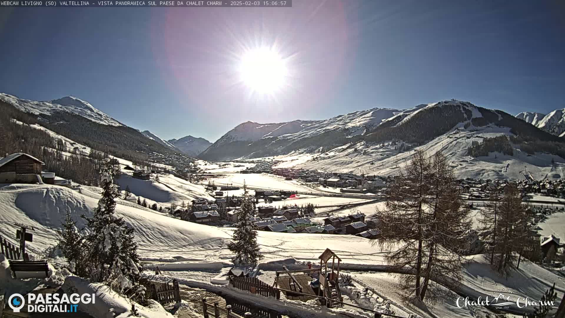 A sunny winter scene shows a snow-covered village nestled in a valley between snow-capped mountains.
