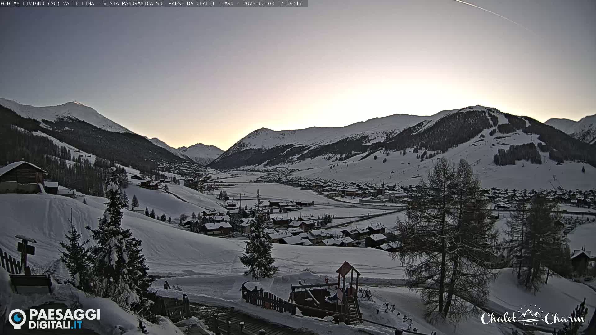 A snow-covered mountain village at dusk, nestled in a valley between snow-capped mountains under a clear, pale sky.