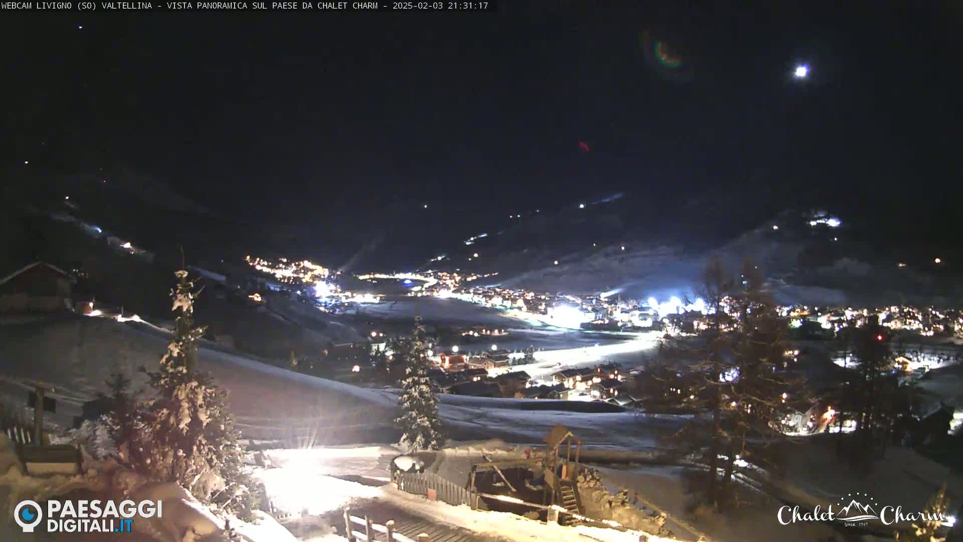 A nighttime, snow-covered mountain village is illuminated by lights, under a dark sky with a visible moon.