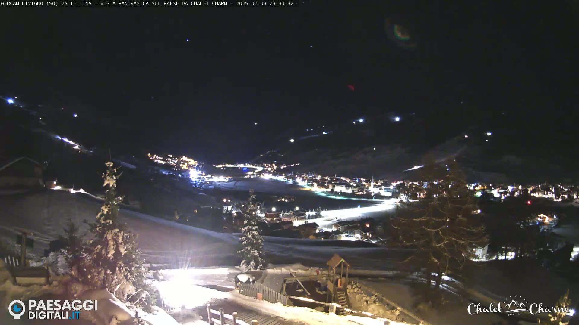 A snow-covered hillside at night overlooks a village illuminated by lights under a clear, dark sky.