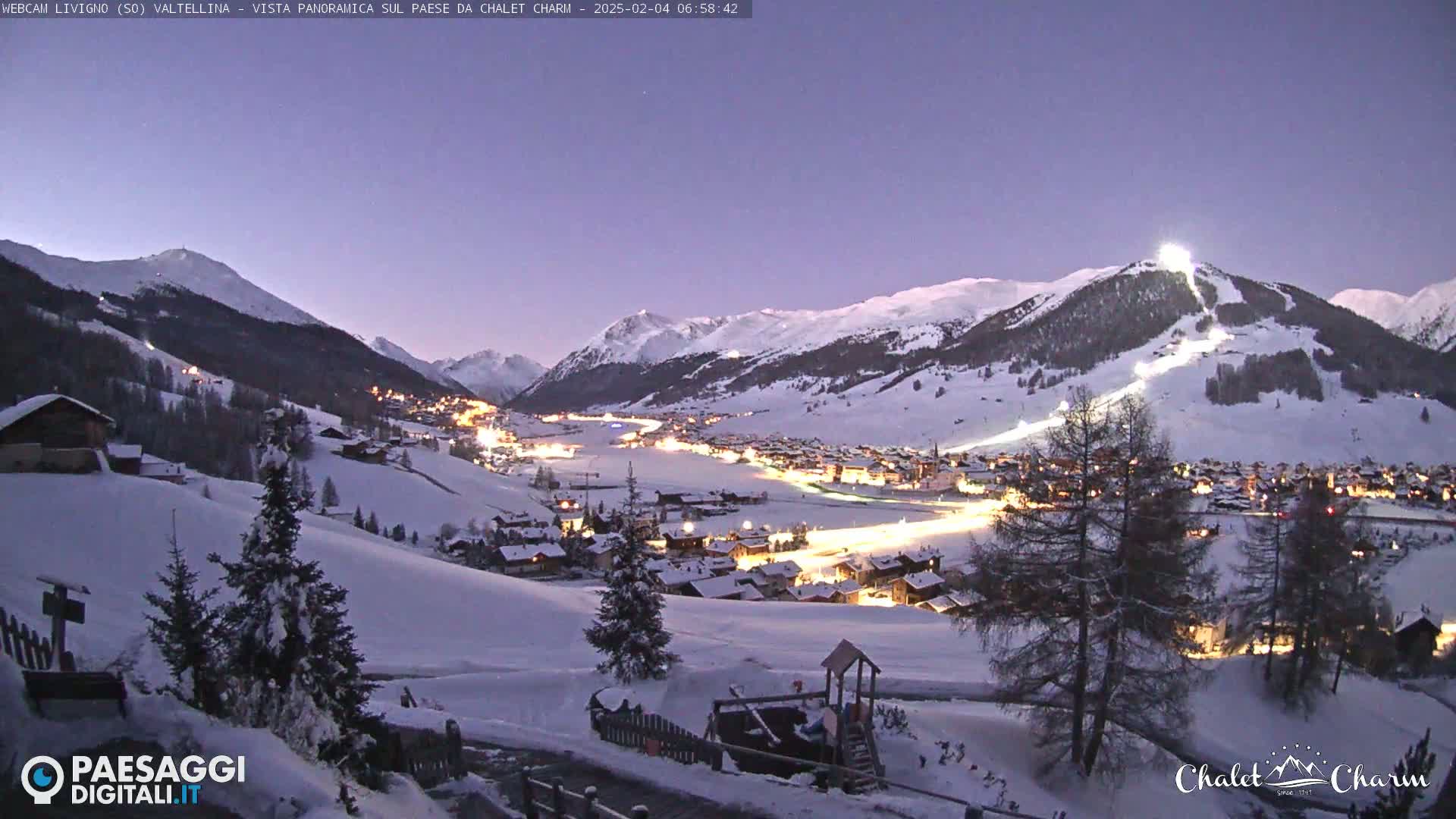 A snow-covered village nestled in a valley between snow-capped mountains is illuminated at twilight.