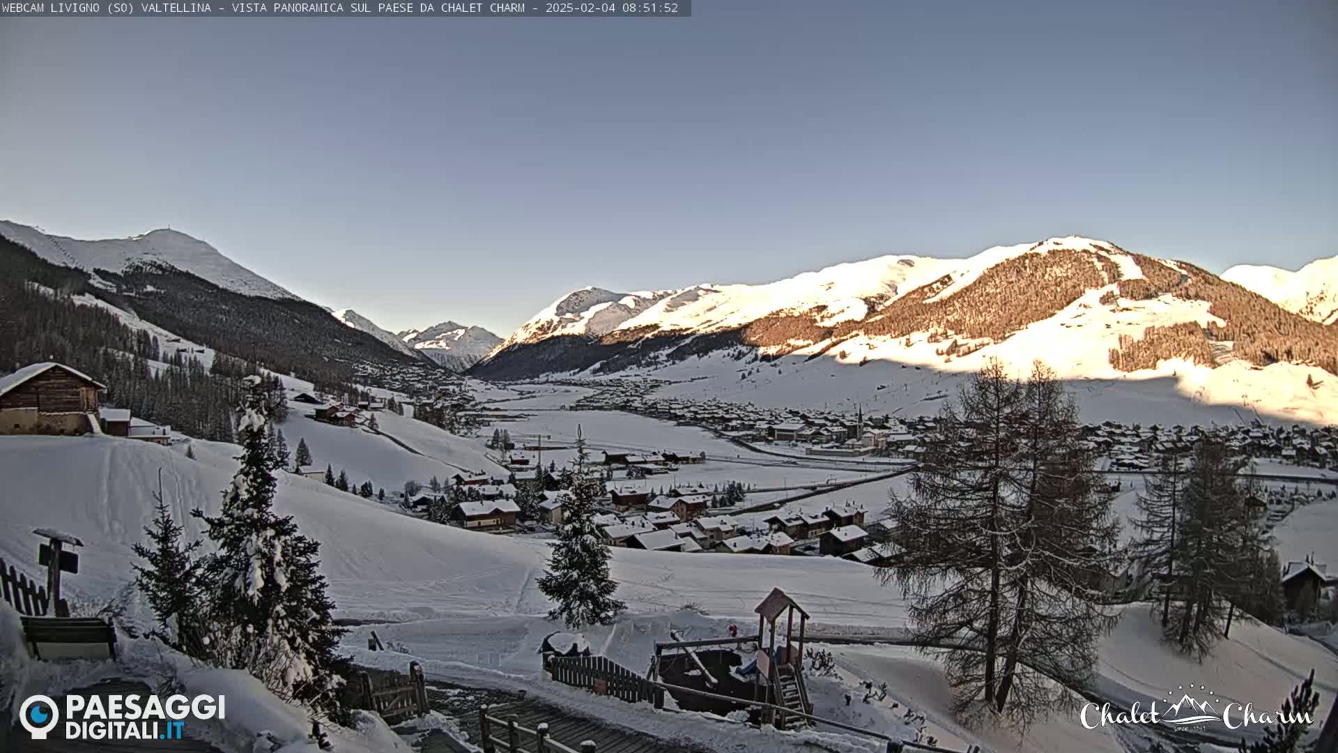 A snow-covered mountain village nestled in a valley beneath sunlit, snow-capped mountains on a clear day.