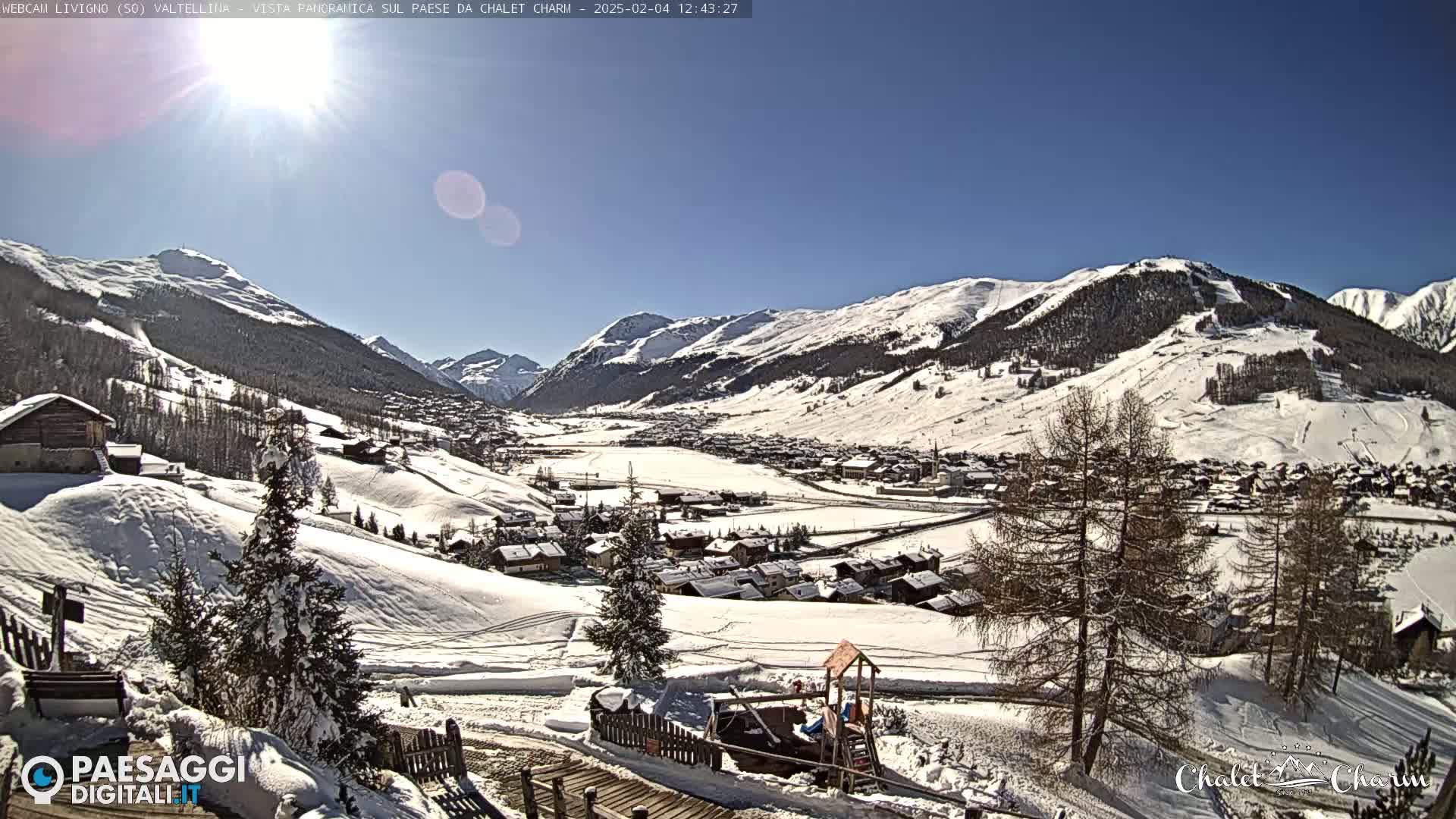A sunny, snow-covered mountain village nestled in a valley, with snow-covered hills and mountains in the background.