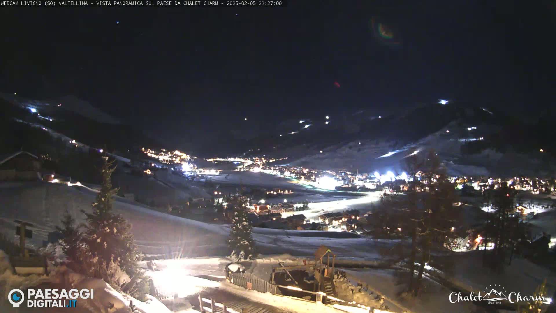A nighttime, snow-covered mountain village is illuminated with lights, seen from a high vantage point under a clear, dark sky.