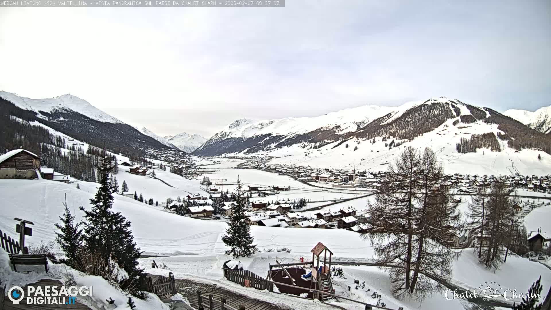 A snow-covered mountain village is nestled in a valley, with overcast skies and snow-covered hills and mountains in the background.