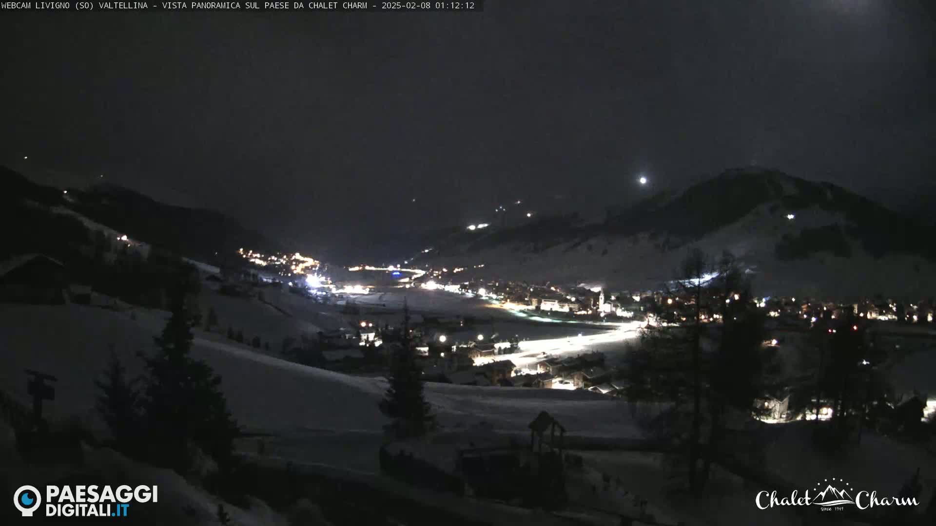 A nighttime, snow-covered mountain village is illuminated by lights, under a dark sky with a visible moon.