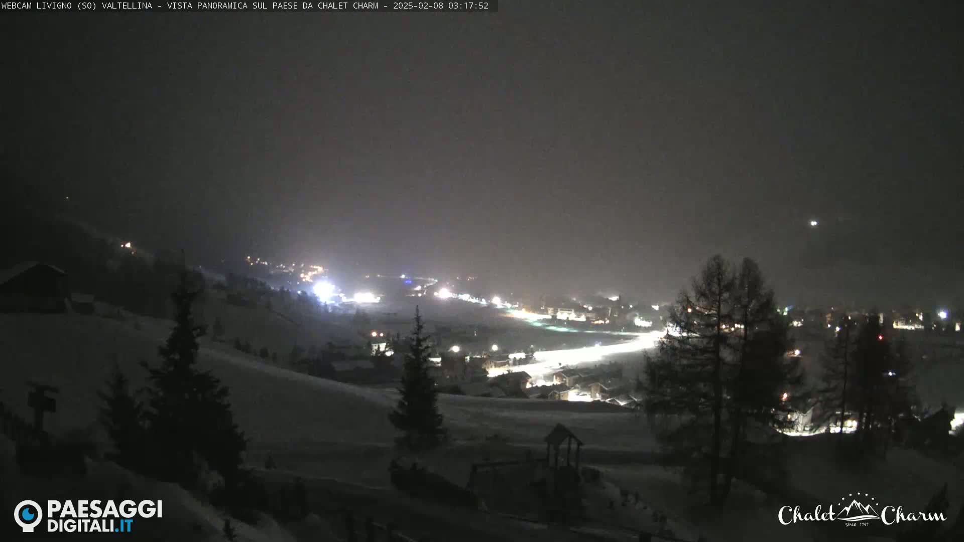 A snow-covered hillside at night overlooks a village illuminated by lights under a dark, cloudy sky.