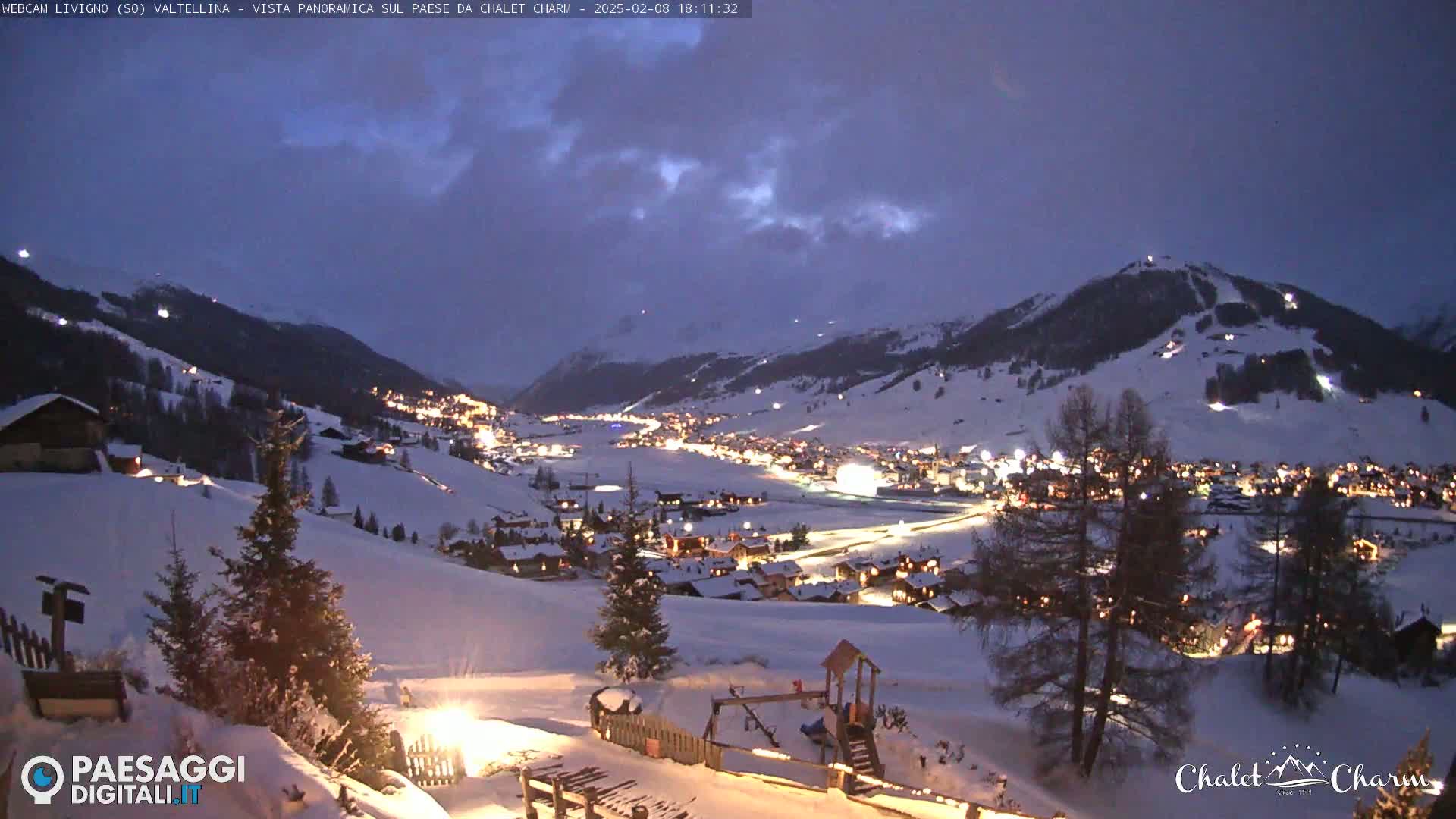 A snow-covered village nestled in a valley at twilight, illuminated by lights, under a partly cloudy sky.