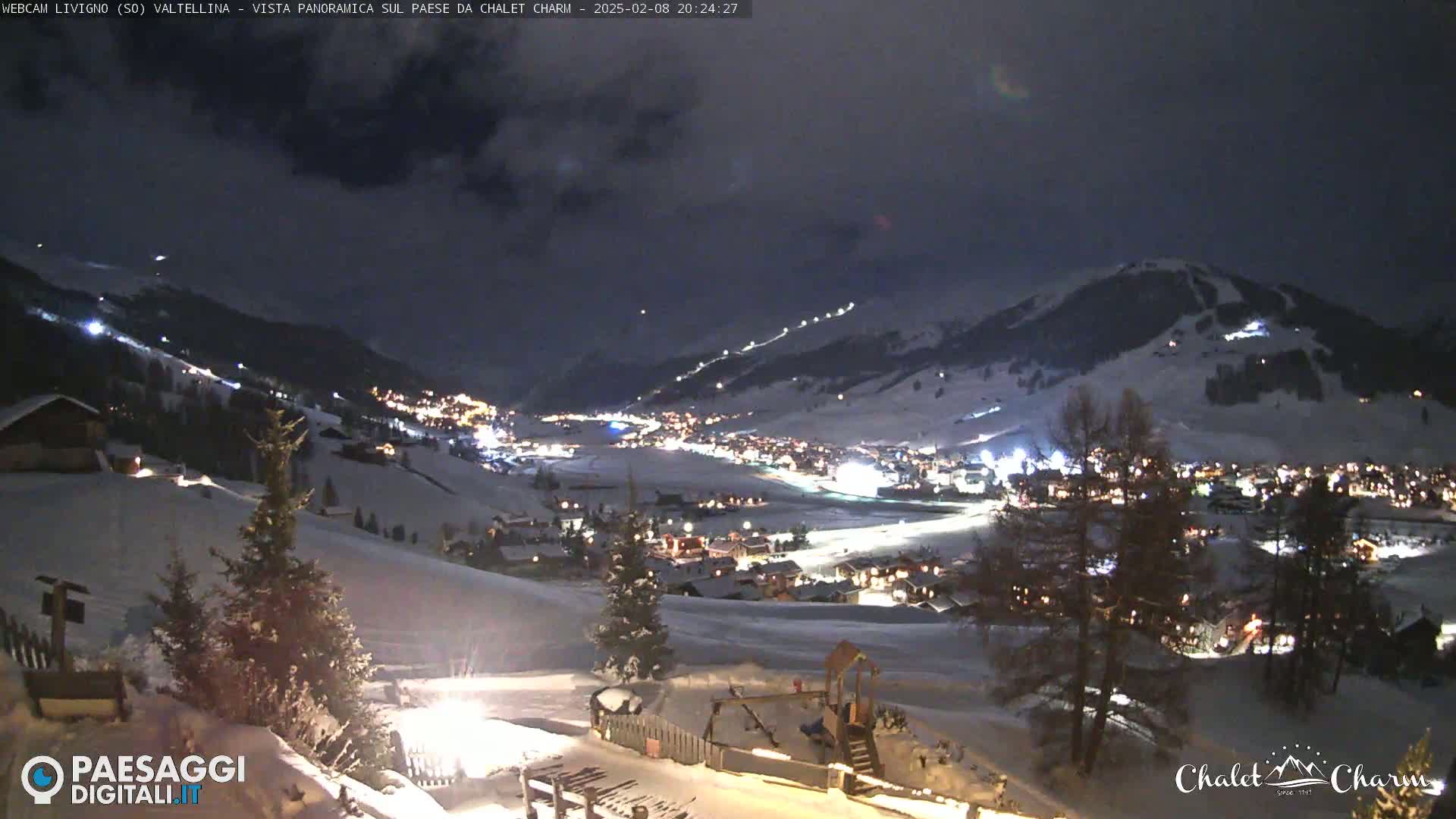 A snow-covered valley town is illuminated at night under a dark, cloudy sky.
