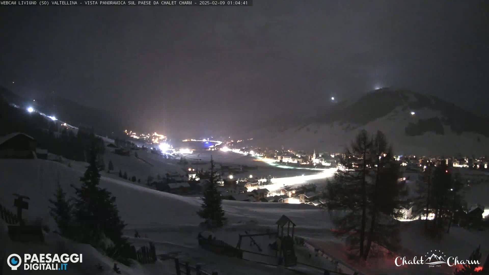 A snow-covered village at night is illuminated by lights, with dark, snow-covered hills in the background under a dark, possibly cloudy sky.