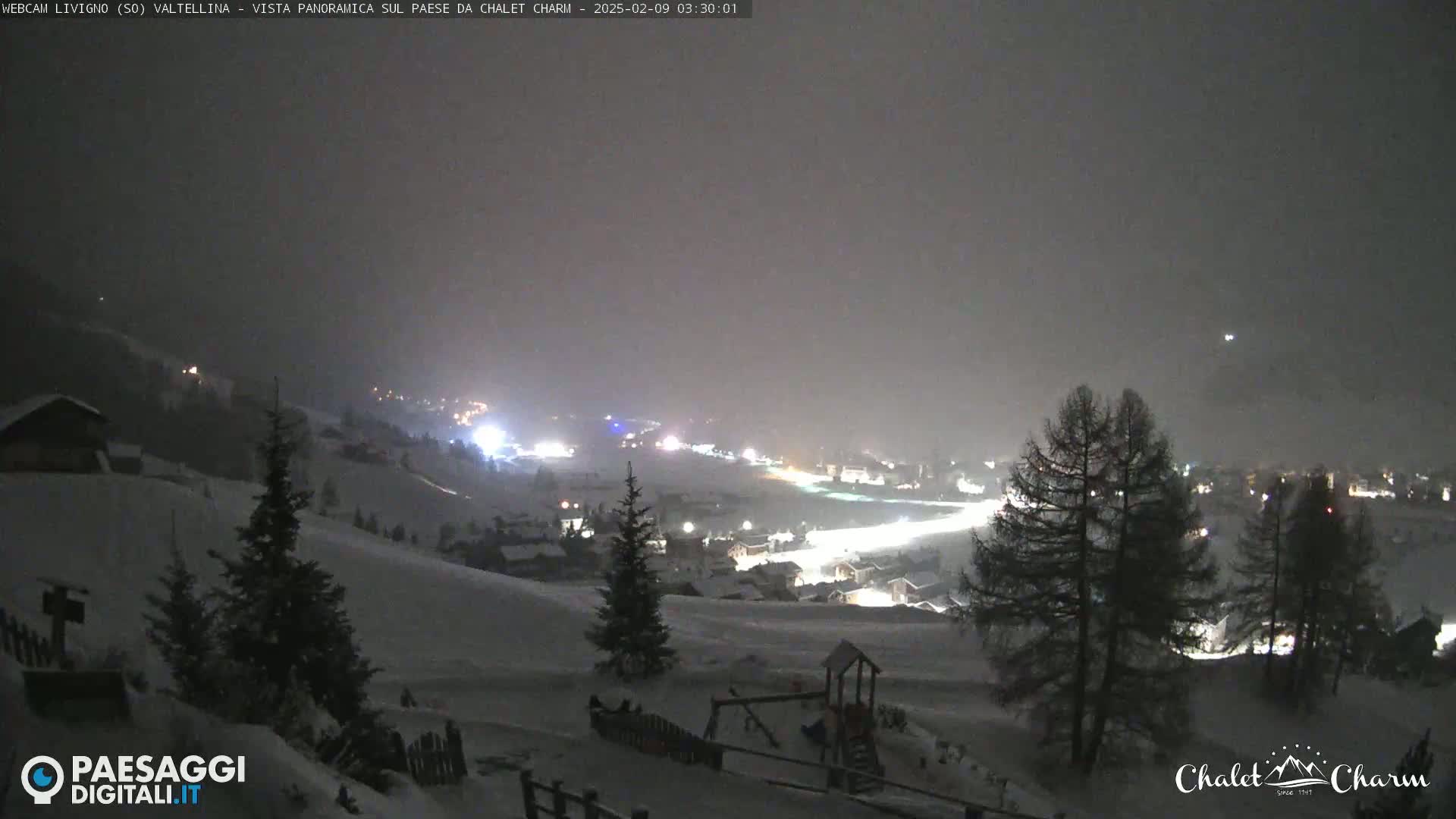 A snowy, nighttime view of a village in a valley, illuminated by lights, under a hazy sky.