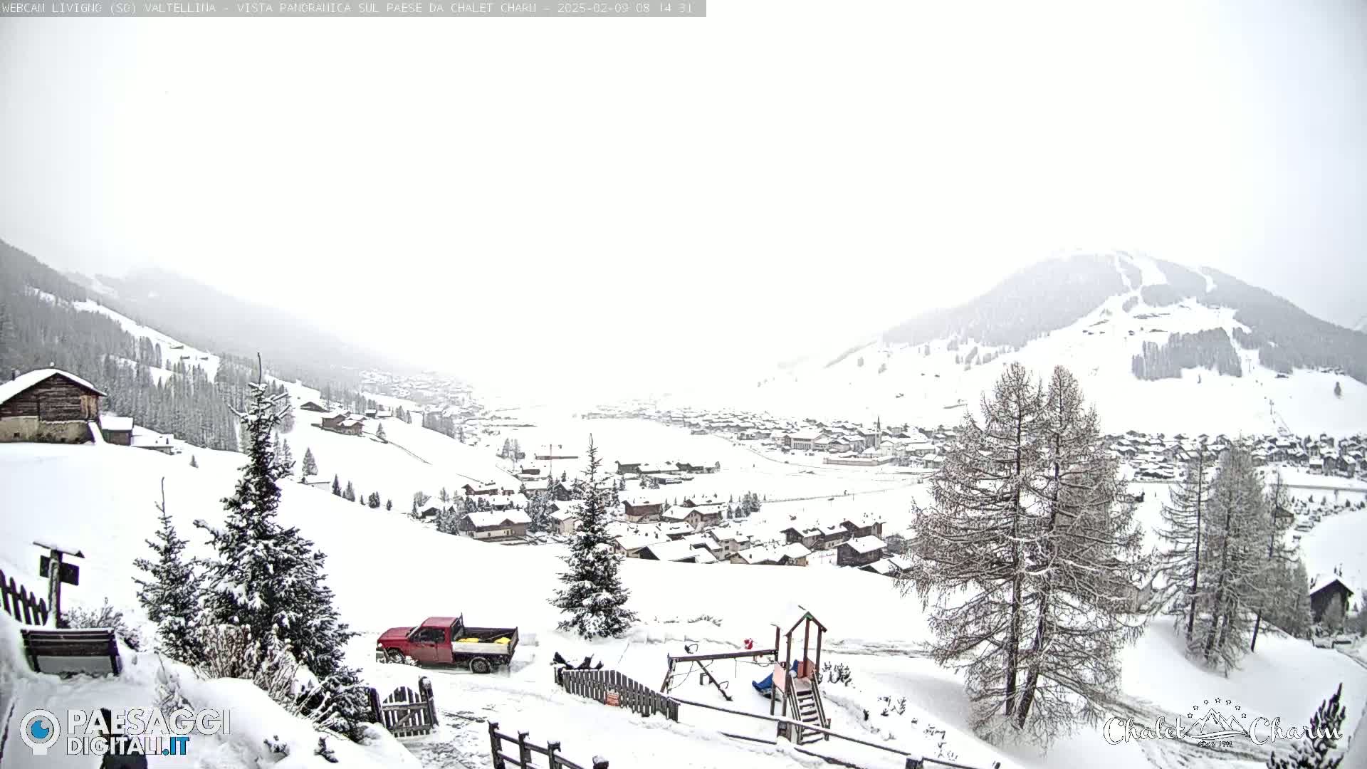 A snowy mountain village is viewed from a hillside, with a red pickup truck parked nearby and a children's playground visible in the foreground under an overcast sky.