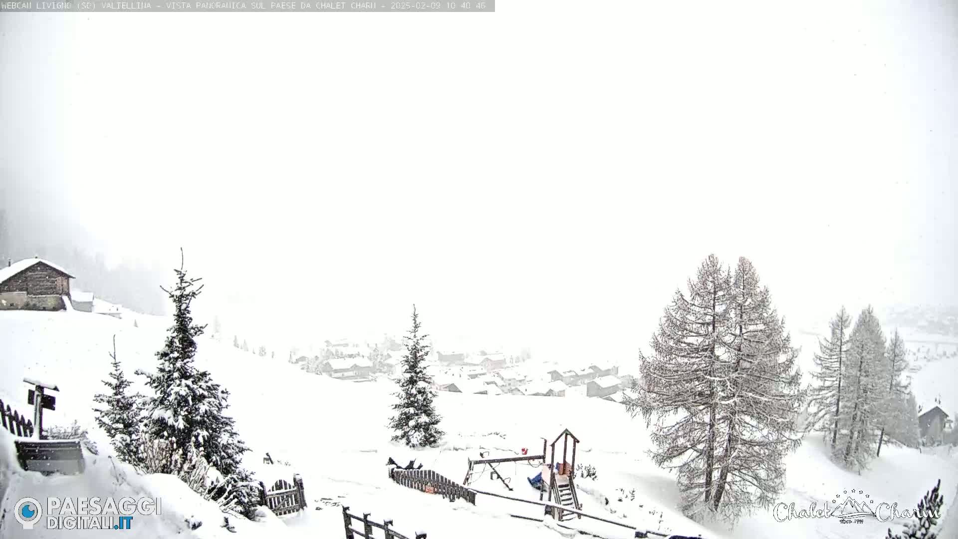 A snow-covered landscape with a small village in the distance, a playground, and several evergreen trees, during a snowfall.