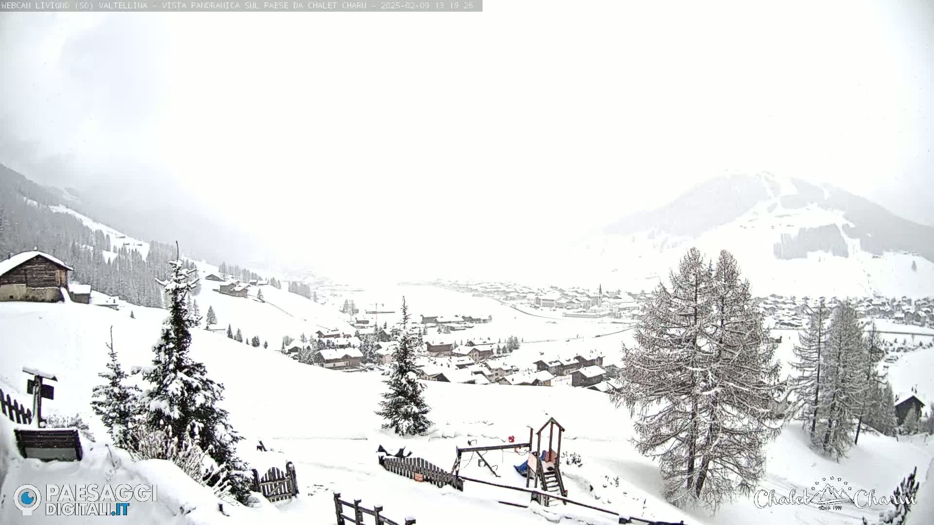 A snow-covered village nestled in a valley below a snow-dusted mountain range, under a heavy, overcast sky.