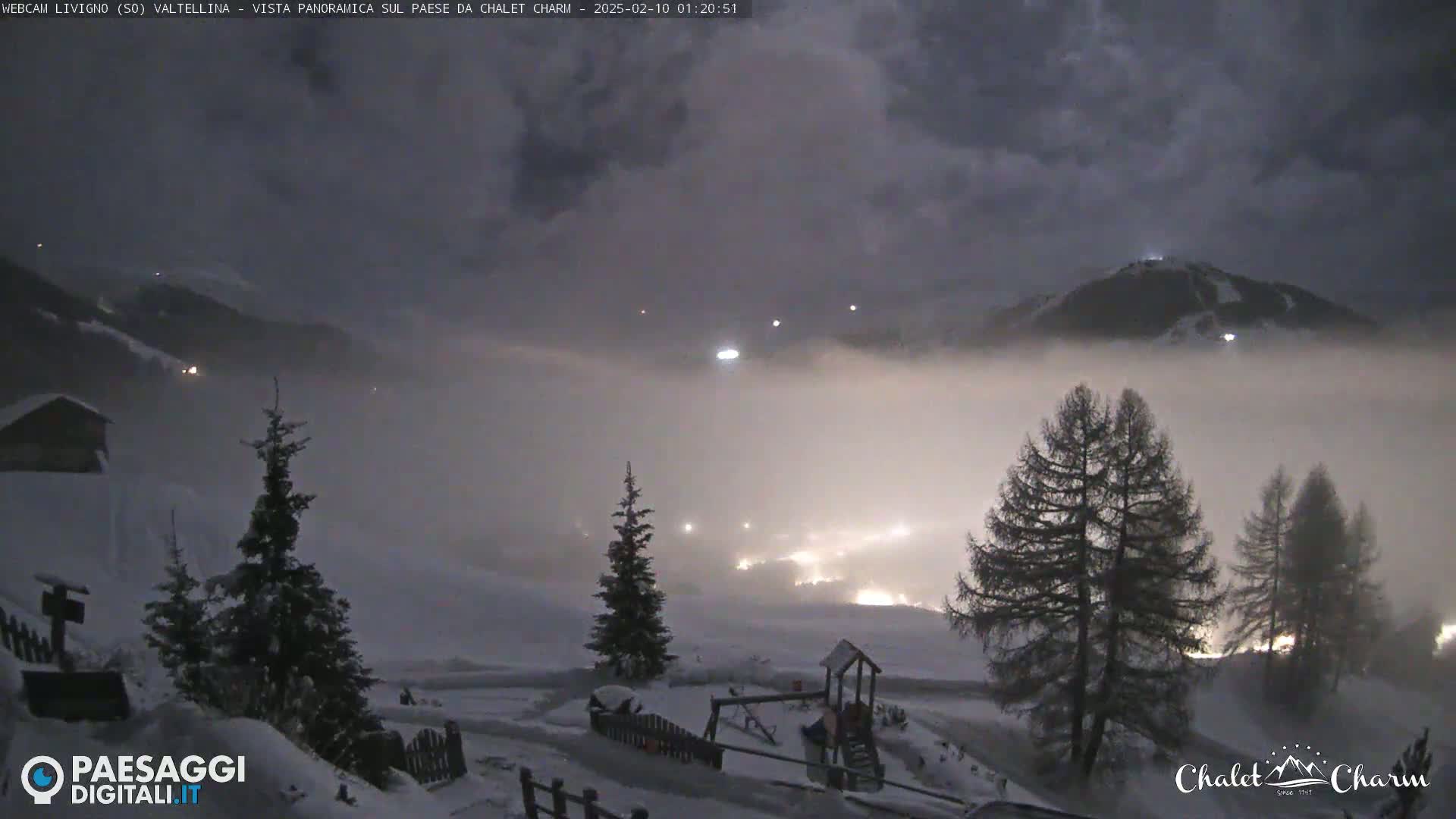A snowy, fog-shrouded mountain landscape at night, illuminated by scattered lights from a town below.