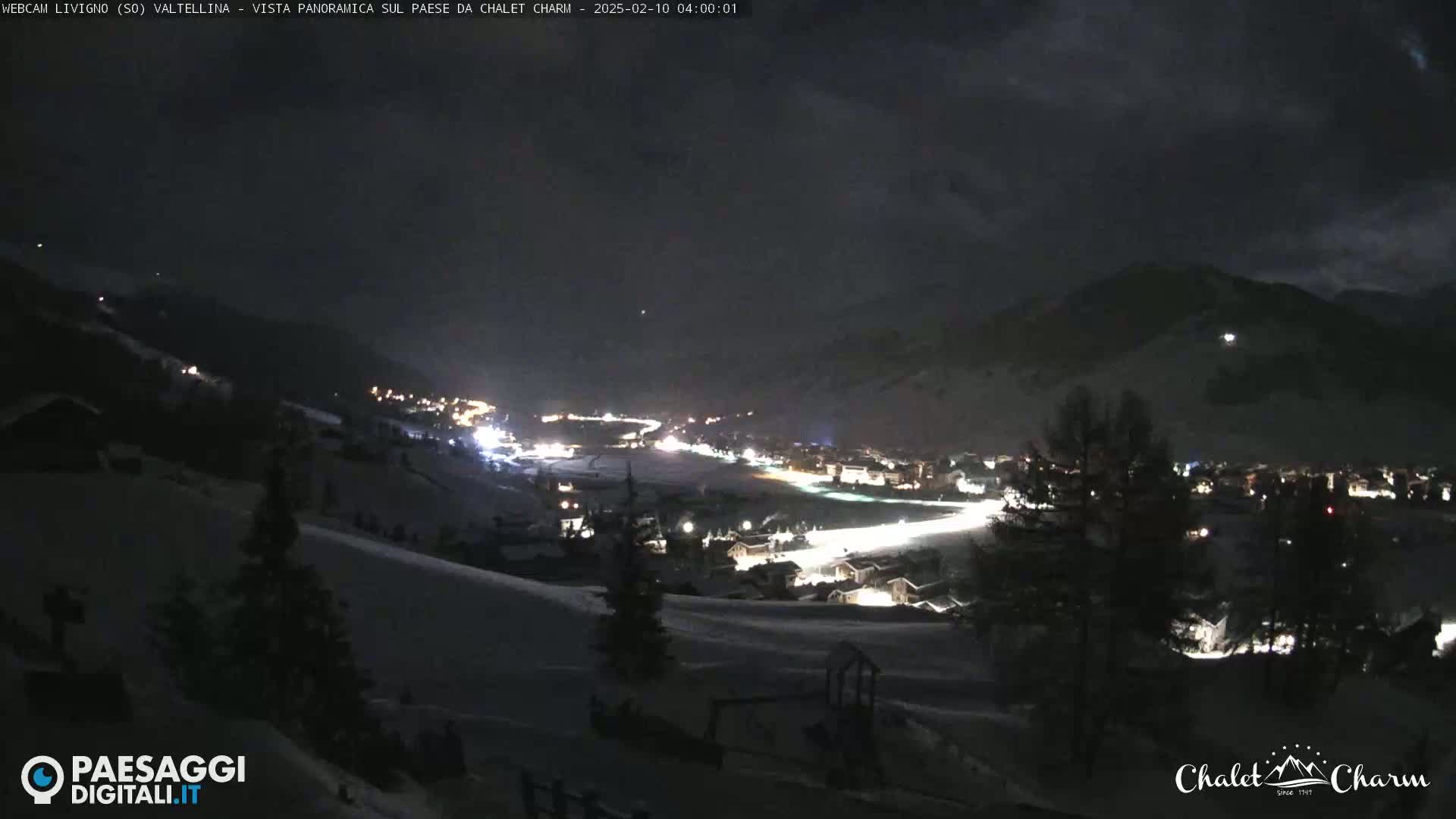 A nighttime panoramic view of a snow-covered town nestled in a valley between mountains under a cloudy sky.