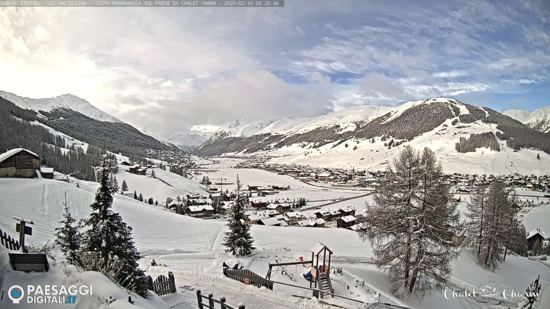 A snow-covered village nestled in a valley between snow-capped mountains under a partly cloudy sky.