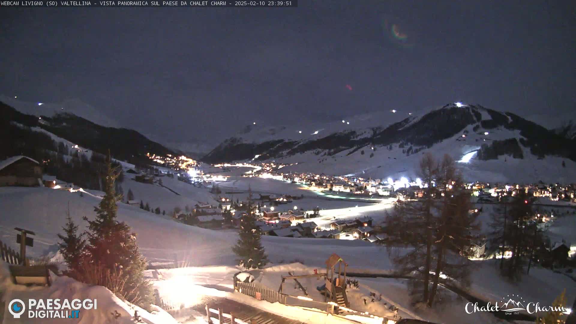 A snow-covered village is illuminated at night, nestled in a valley between snow-capped mountains under a dark sky.