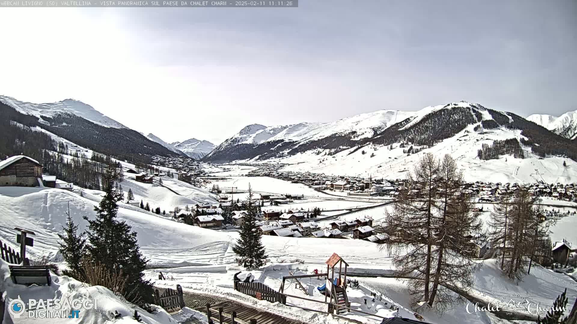 A snow-covered village nestled in a valley between snow-capped mountains on a partly cloudy day.