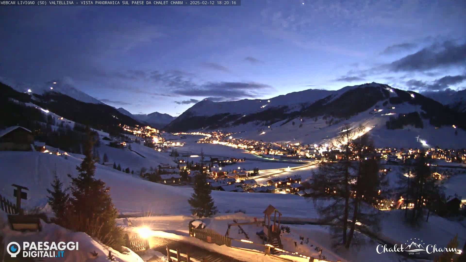A nighttime, panoramic view of a snow-covered village nestled in a valley between illuminated ski slopes and mountains under a twilight sky.