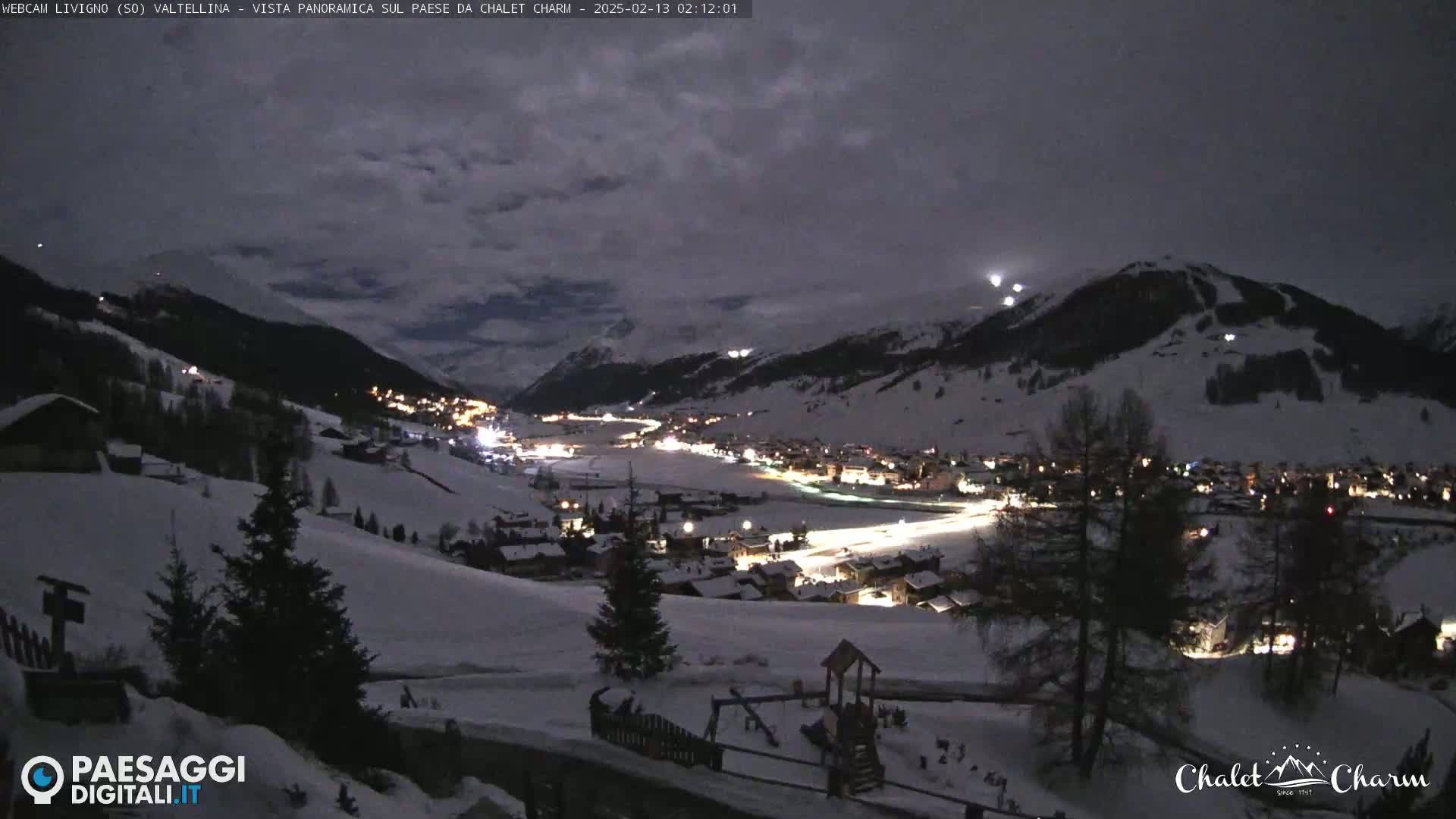 A snow-covered village nestled in a valley at night under a cloudy sky, illuminated by lights from buildings and possibly moonlight.