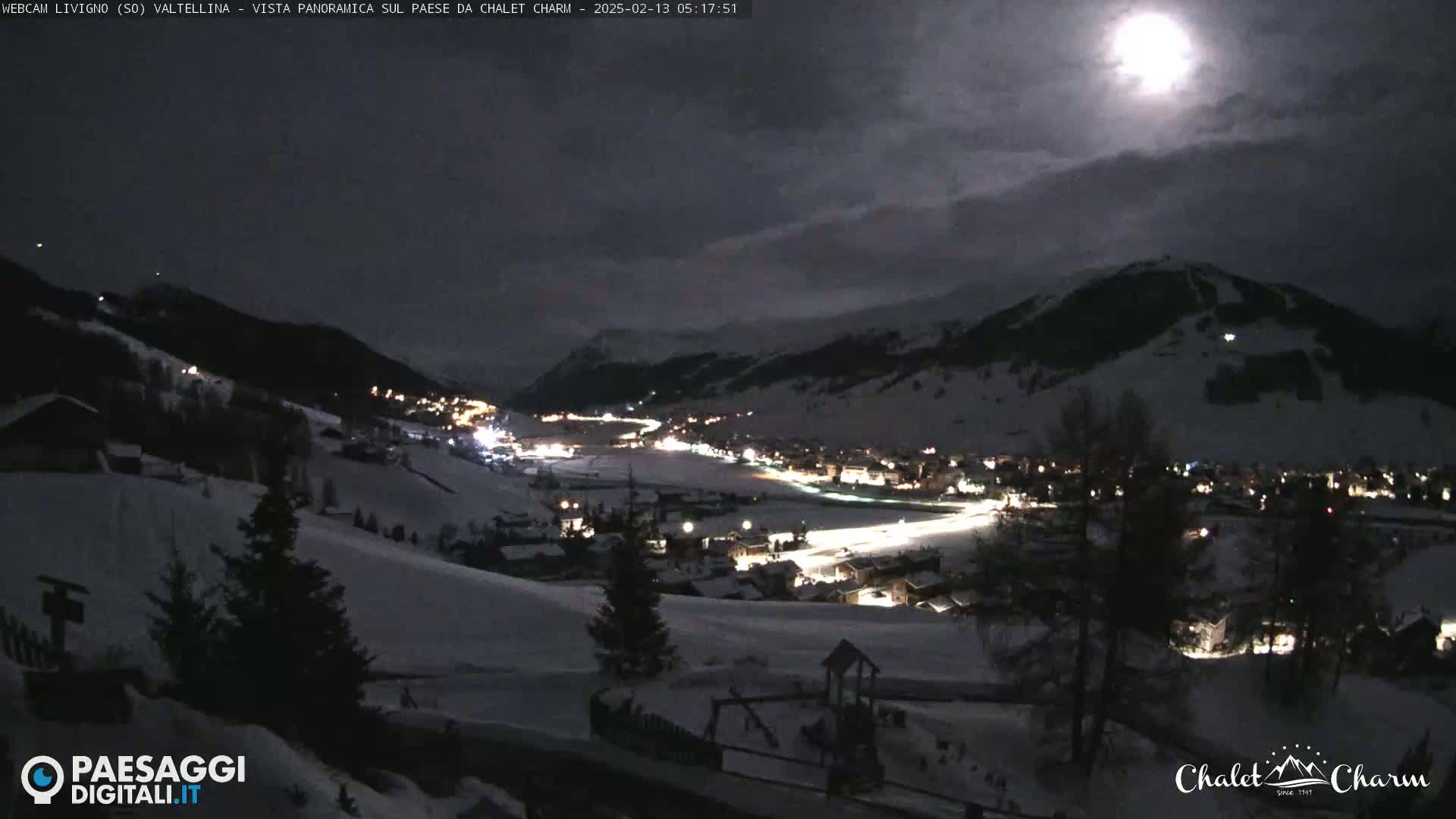A moonlit, snow-covered mountain village is seen at night, under a partly cloudy sky.