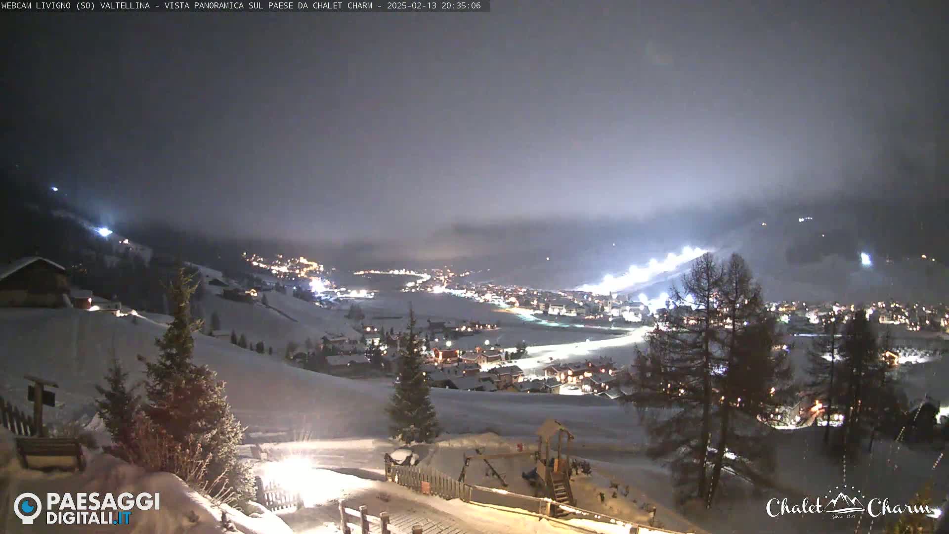 A nighttime, panoramic view of a snow-covered village illuminated by lights, under a cloudy sky.