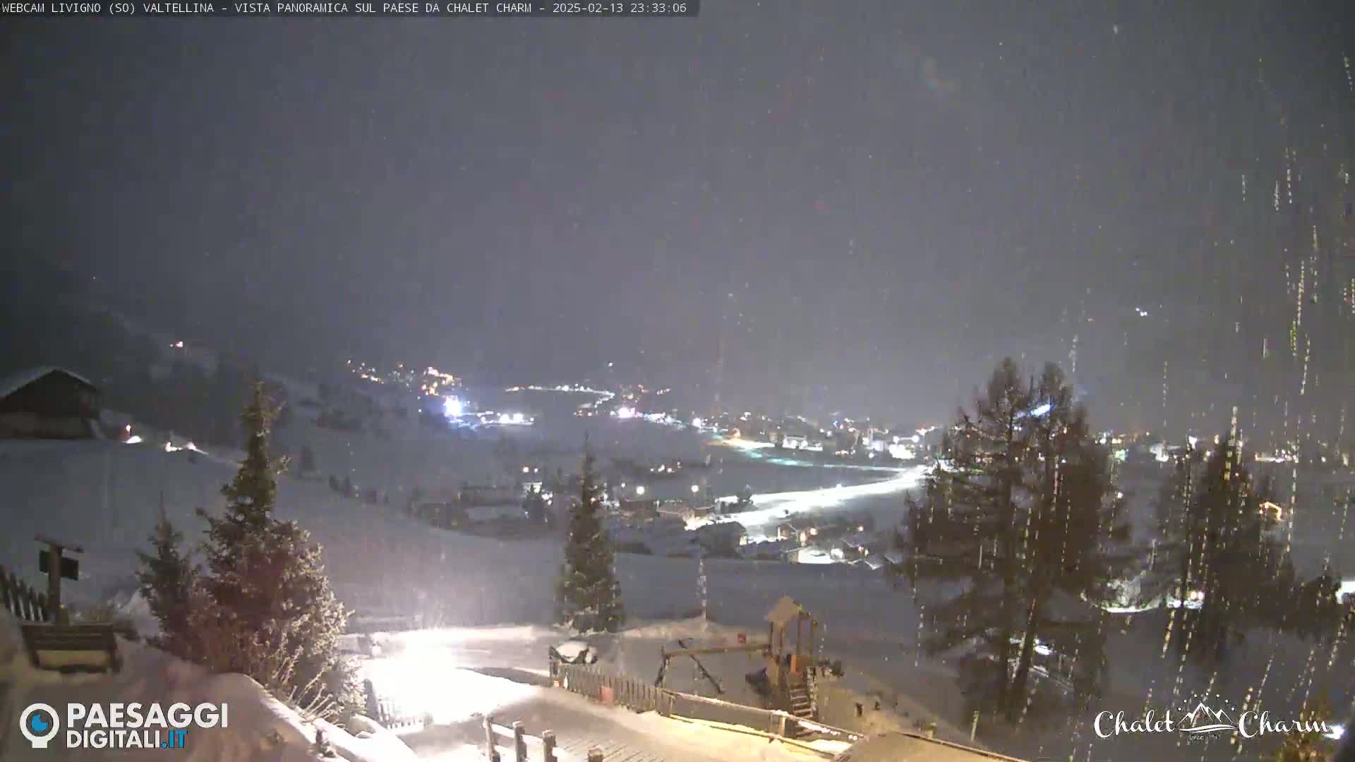 A snow-covered landscape at night, viewed from a hilltop, shows a village with lights illuminated below, under a falling snow.