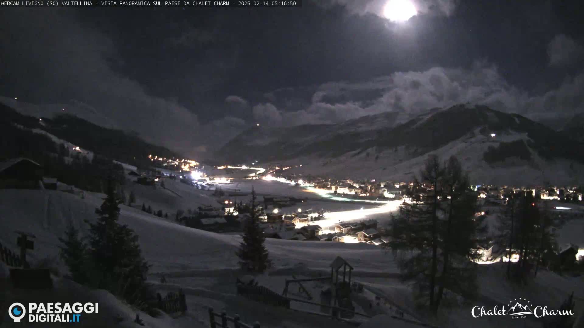 A nighttime, snowy mountain village scene is illuminated by a bright moon partially obscured by clouds.