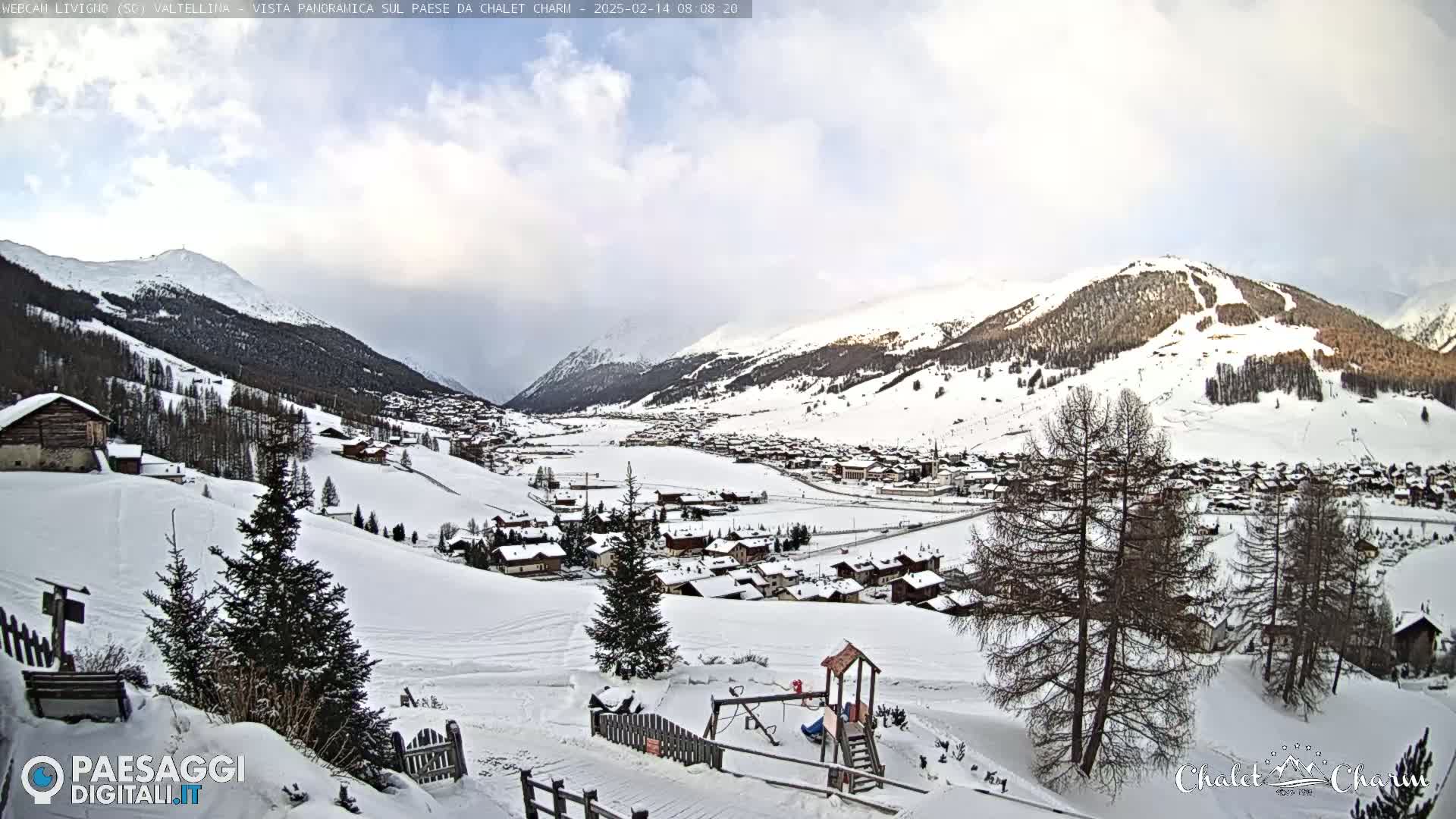A snow-covered village nestled in a valley between snow-capped mountains under a partly cloudy sky.