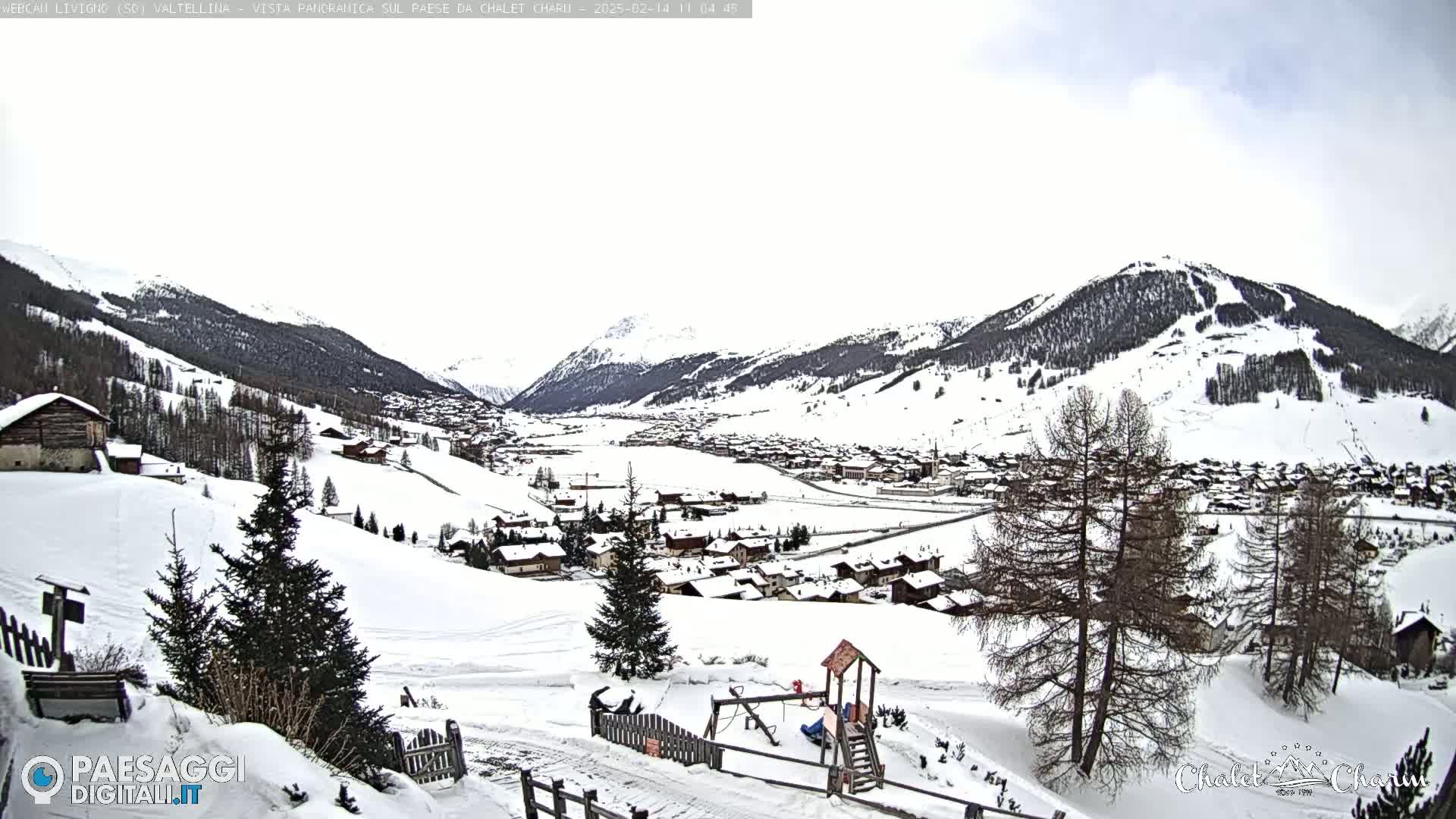 A snow-covered village nestled in a valley between snow-capped mountains under an overcast sky.