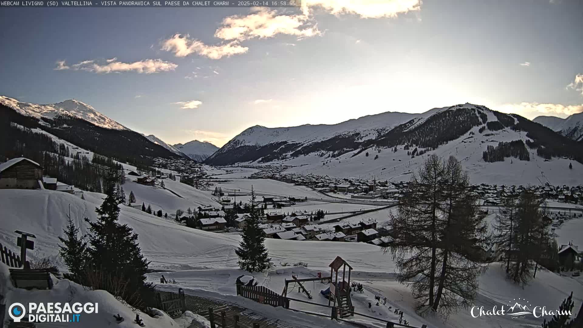 A snow-covered village nestled in a valley between snow-capped mountains under a partly cloudy sky.