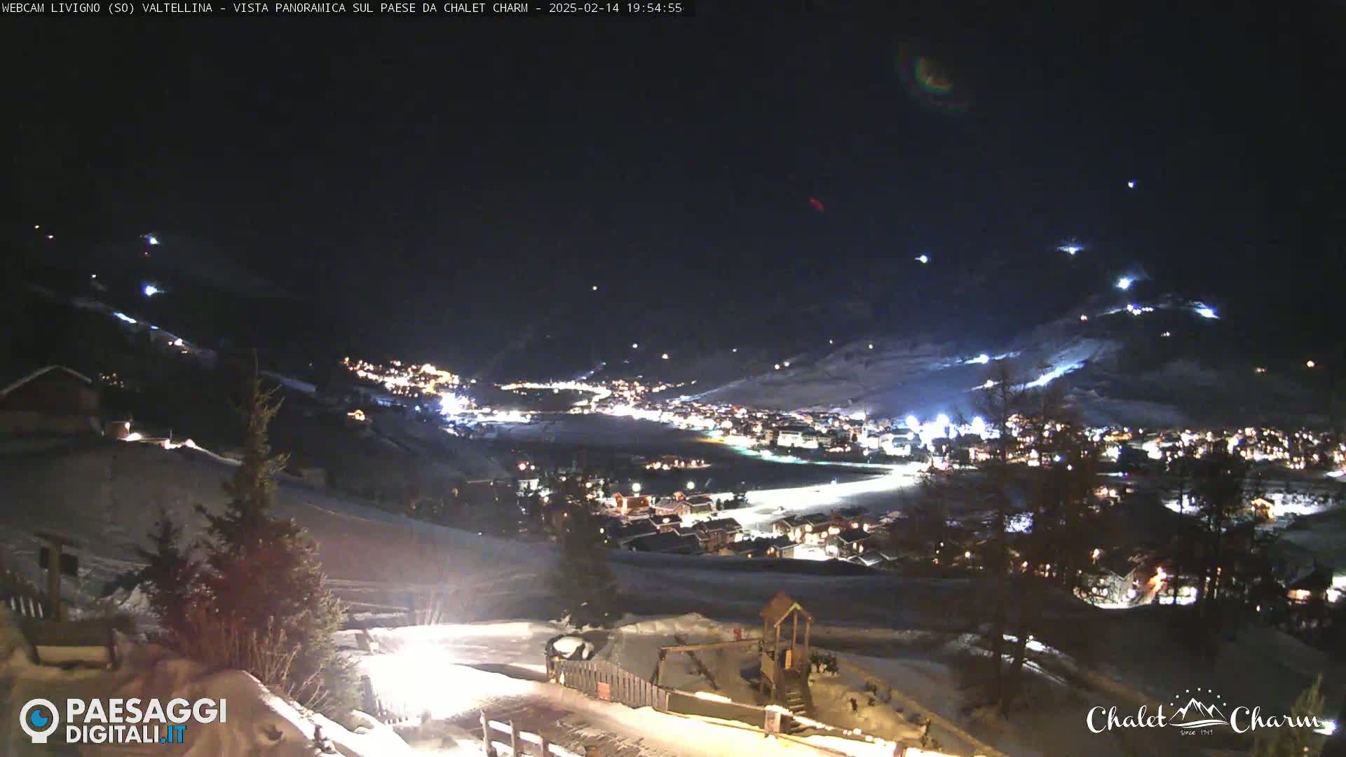 A nighttime, panoramic view of a snow-covered town nestled in a valley, illuminated by lights, under a dark sky.