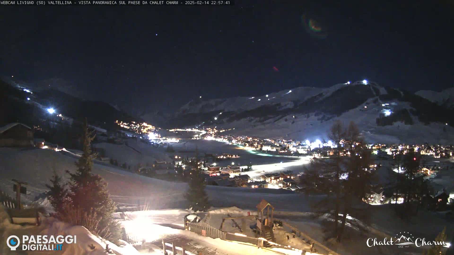A nighttime, snowy panorama shows a village nestled in a valley illuminated by lights, with snow-covered hills and mountains in the background under a dark, clear sky.