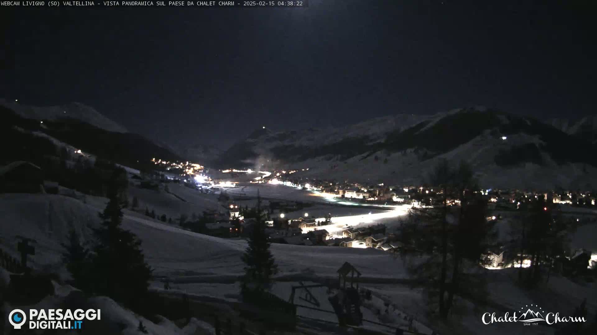 A snow-covered valley at night, illuminated by the lights of a village nestled among dark mountains under a moonlit sky.