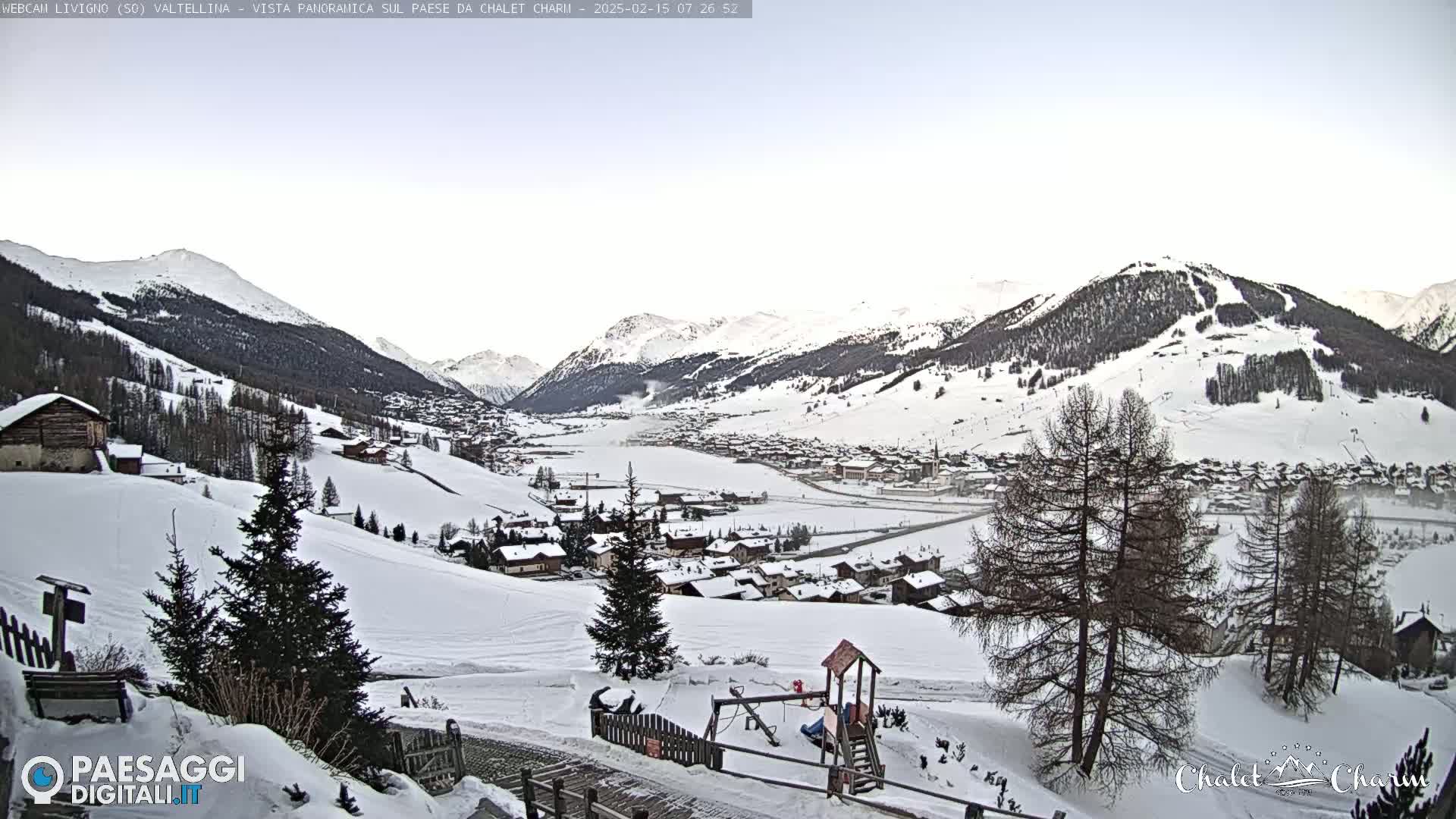 A snow-covered village nestled in a valley between snow-capped mountains under a clear, pale sky.