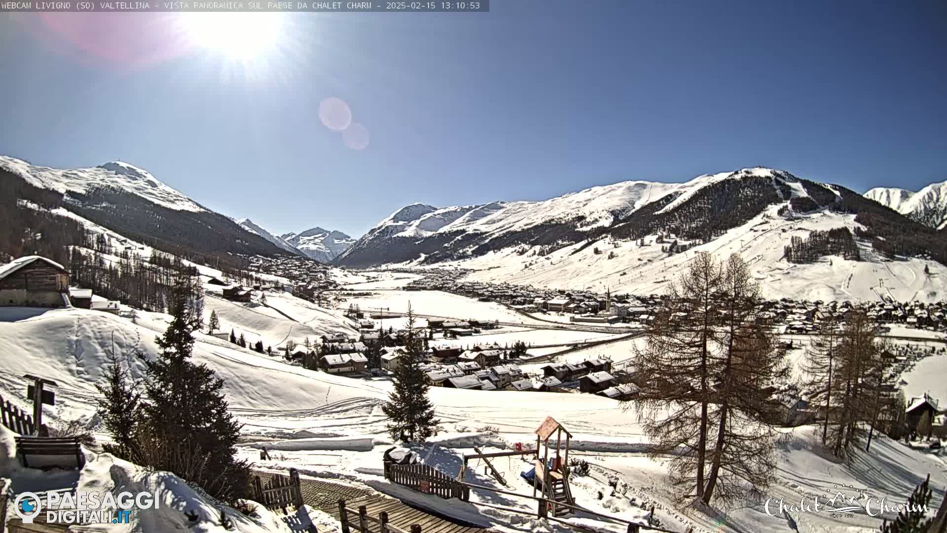 A sunny, snow-covered mountain village nestled in a valley, with surrounding hills also covered in snow.