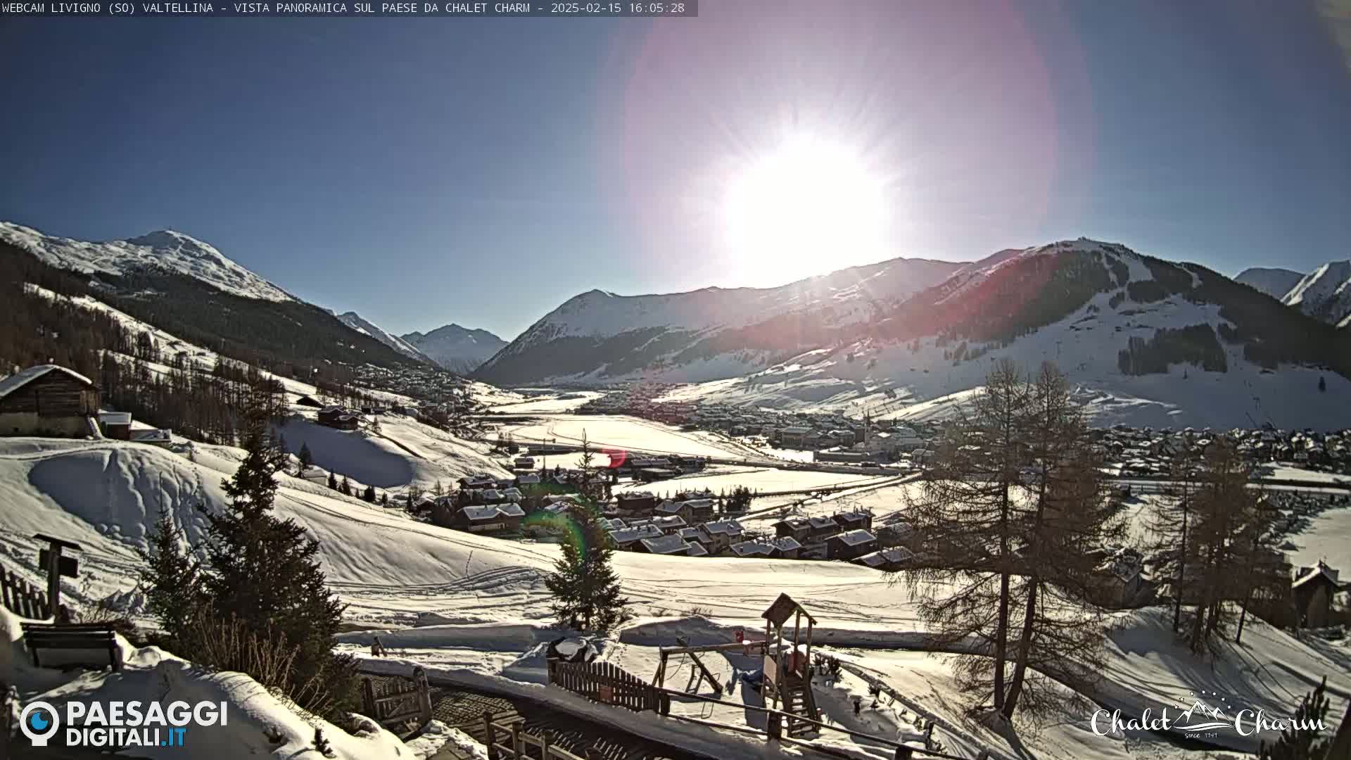 A sunny, snow-covered mountain valley village is viewed from a high vantage point, with a playground visible in the foreground.