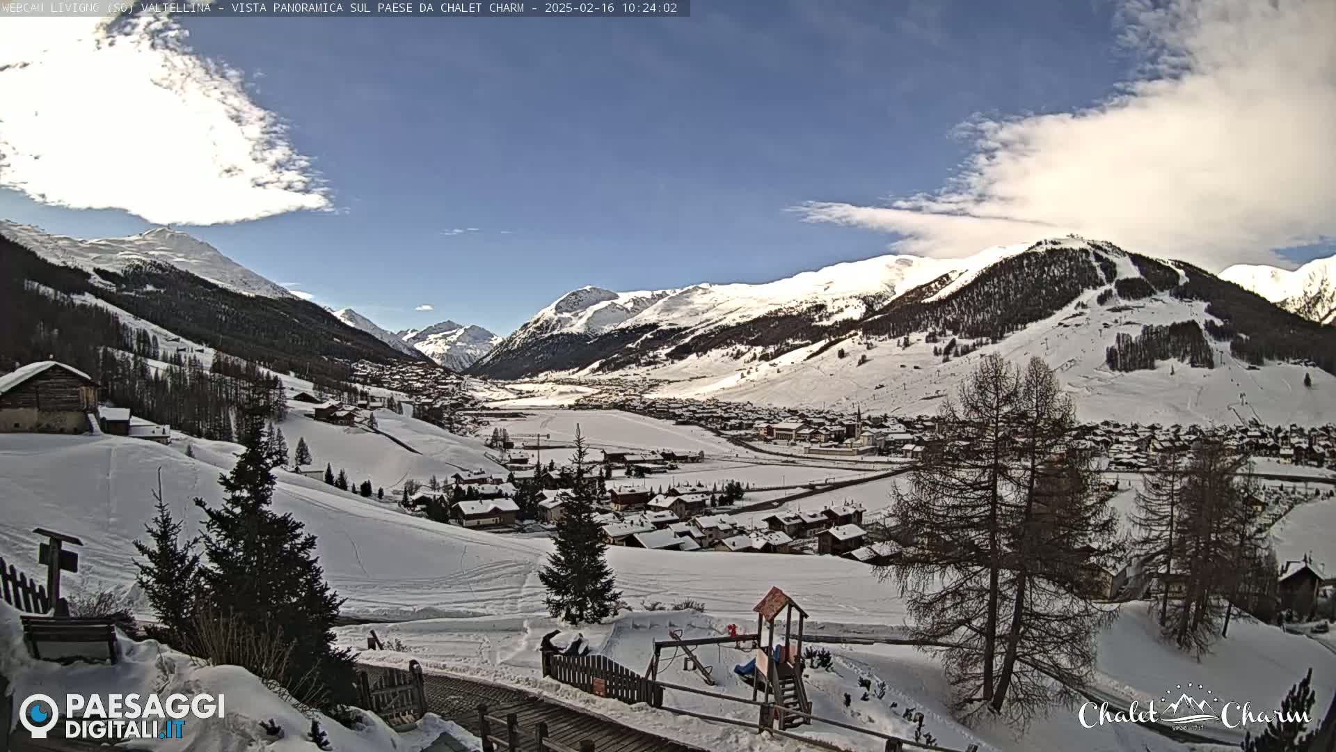 A snow-covered village nestled in a valley between snow-capped mountains under a mostly sunny sky with some scattered clouds.