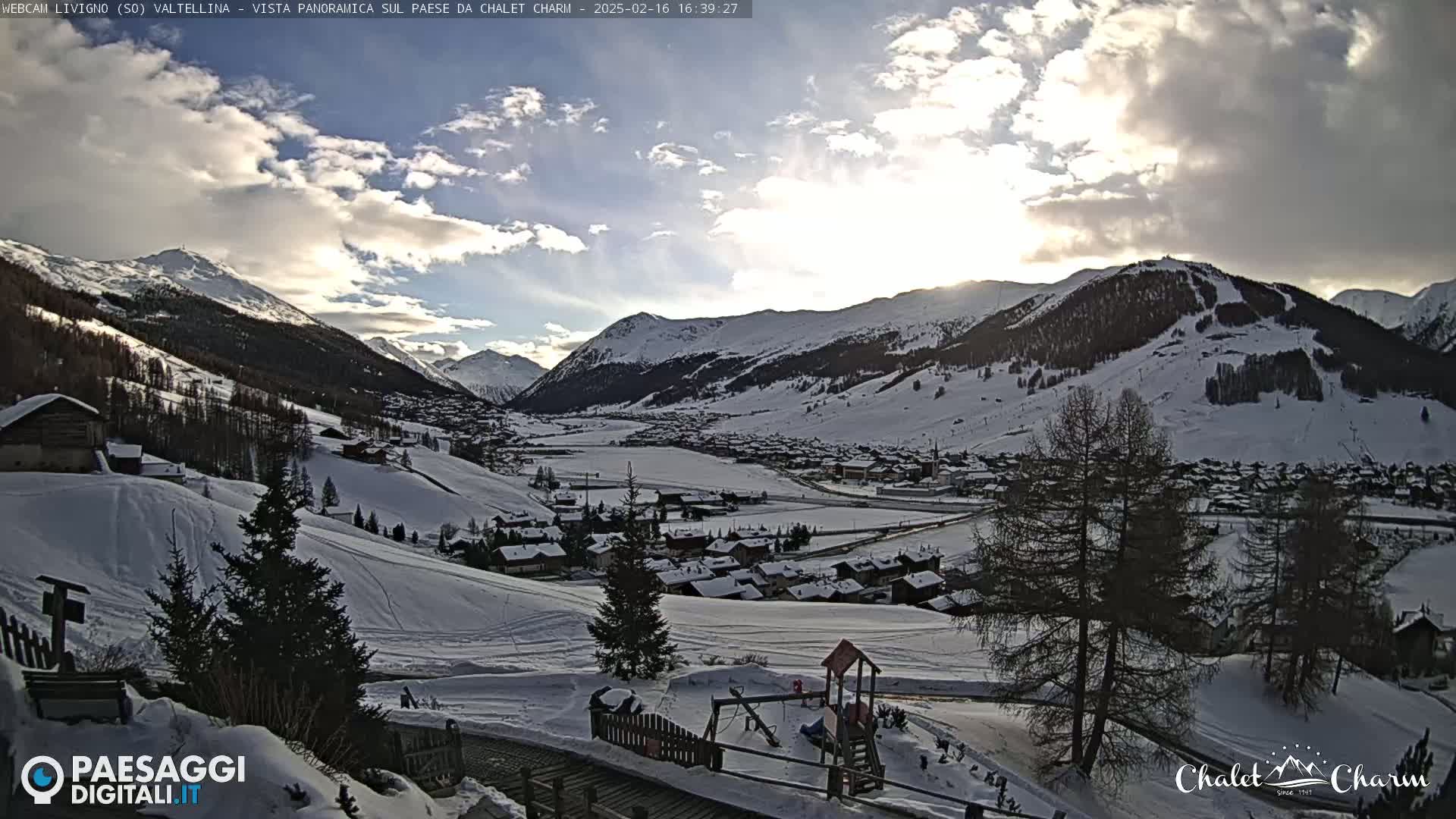 A snow-covered valley town is nestled among partially cloudy mountains under a partly sunny sky.