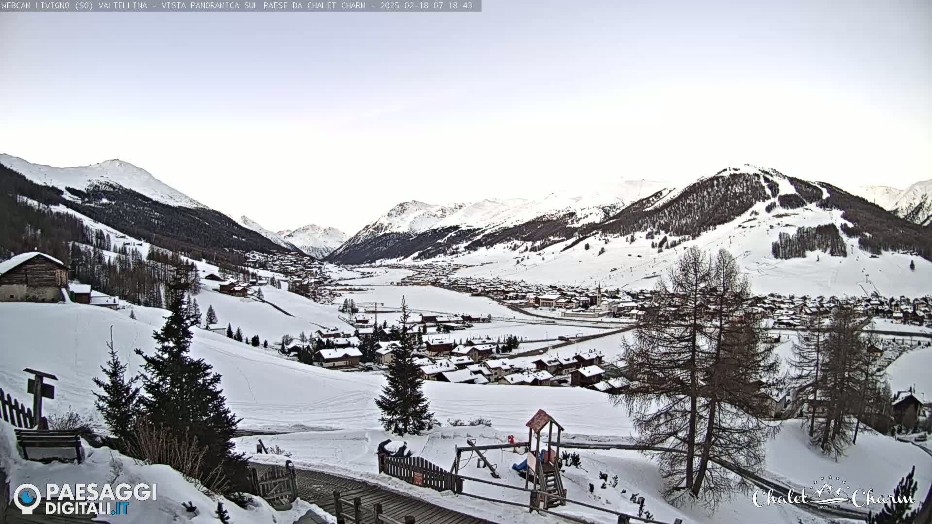 A snow-covered village nestled in a valley between snow-capped mountains under a clear, pale sky.